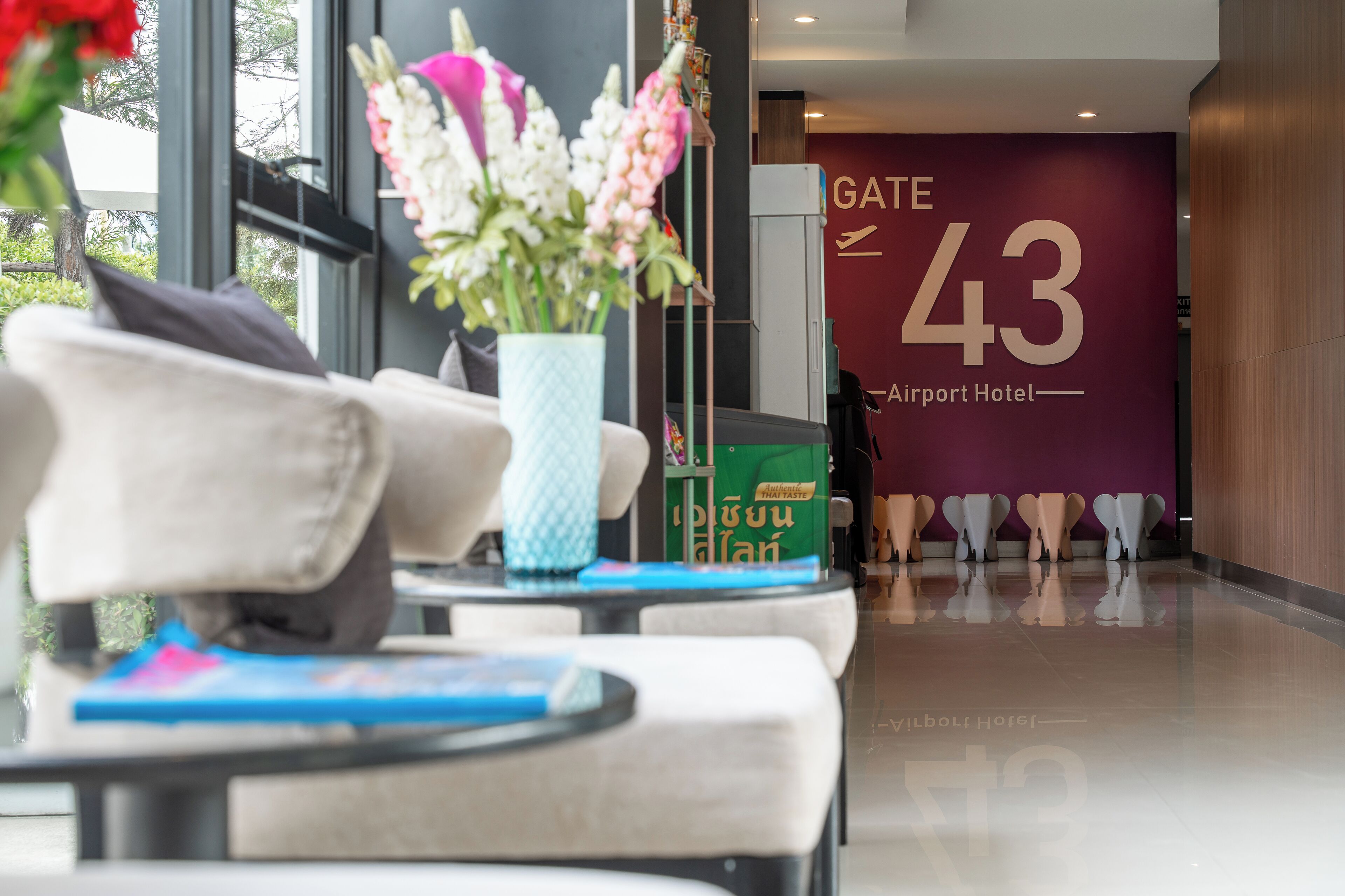 A hotel lobby with a reception desk, a large sign reading "GATE 43 Airport Hotel," and a view of the outside through large windows.