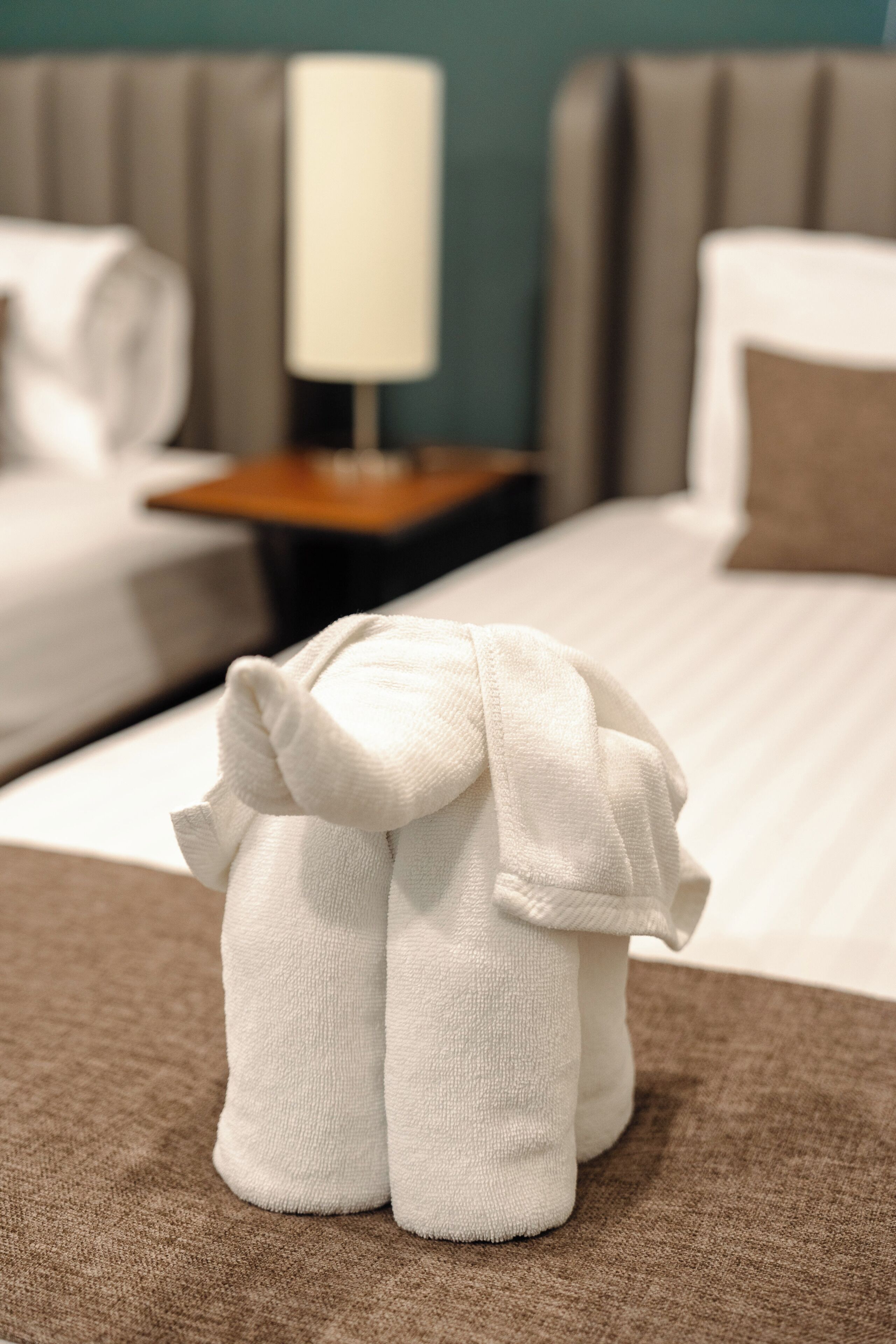 Een net opgemaakt bed met een kussen, een nachtkastje met een lamp en een handdoeksculptuur op het bed.
