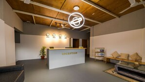 Uma área de recepção com balcão de atendimento identificado como "RECEPTION", um relógio, uma planta, um banco e um ventilador de teto.