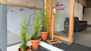 Entrada de hotel com porta de madeira, placa com a inscrição "Shine Residency" e duas plantas em vasos sobre um tapete preto.