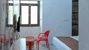 A compact room with a bed, a small table, a red chair, a ceiling fan, and a window. - Hotel Apni Haveli Villa (Udaipur)
