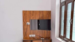 A compact room with a wooden wall unit, a TV, a small fridge, a red chair, a red table, and a cylindrical stool. - Hotel Apni Haveli Villa (Udaipur)
