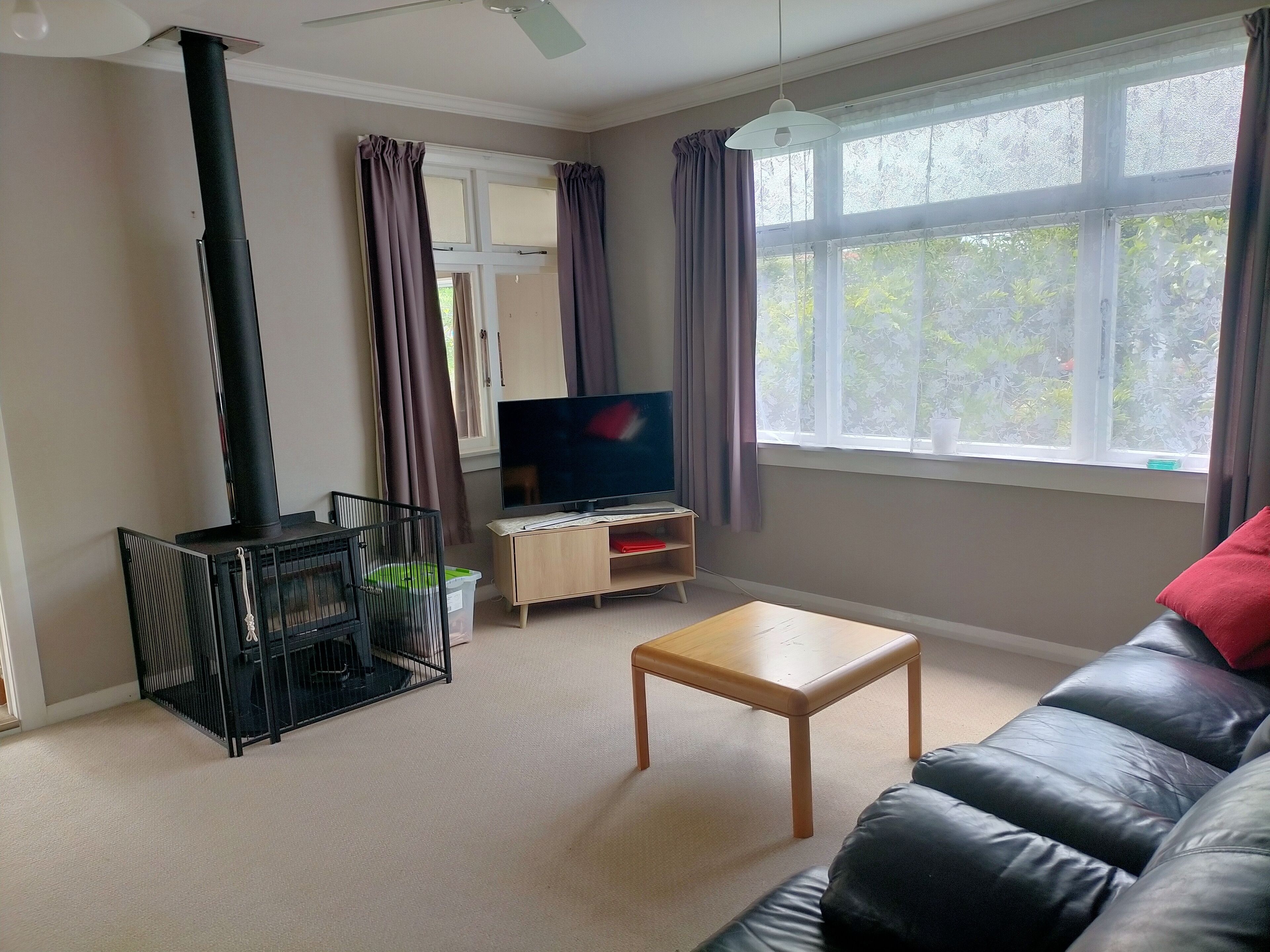A living room with a black leather sofa, a wooden coffee table, a TV on a wooden stand, a black grill, and a window with curtains.