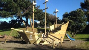 Une structure de jeu en forme de bateau de pirate en bois, installée dans un parc et comprenant un toboggan et un filet d’escalade.