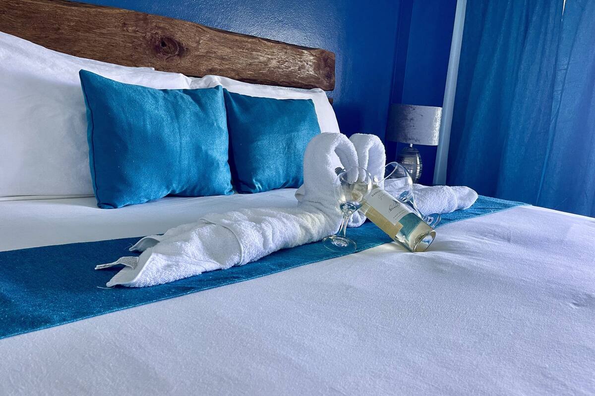 A neatly made bed with a towel and a bottle of wine, blue pillows, and a wooden headboard.