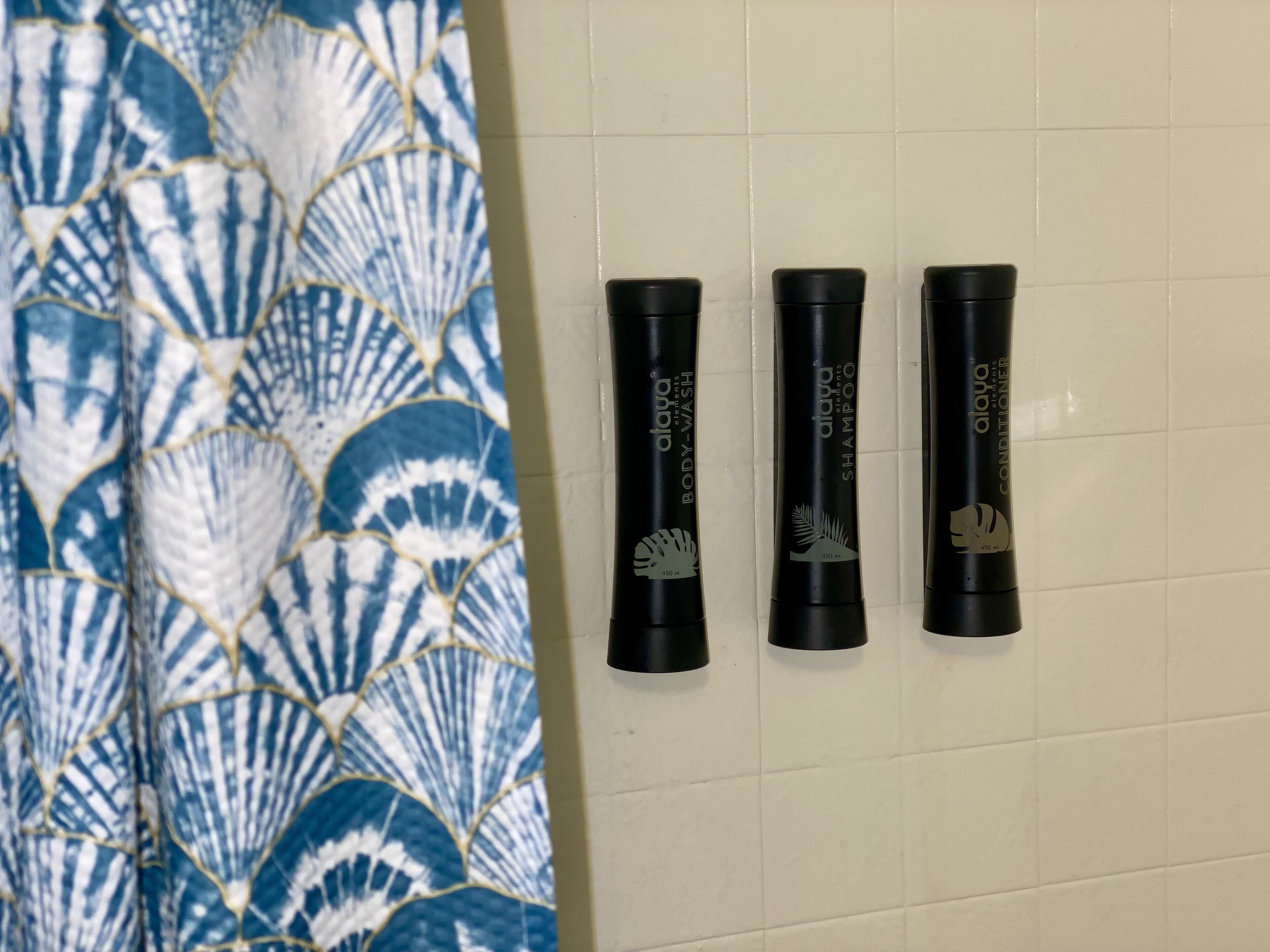 Trois flacons noirs de gel douche et d’après-shampoing Alapa, posés sur un mur carrelé.