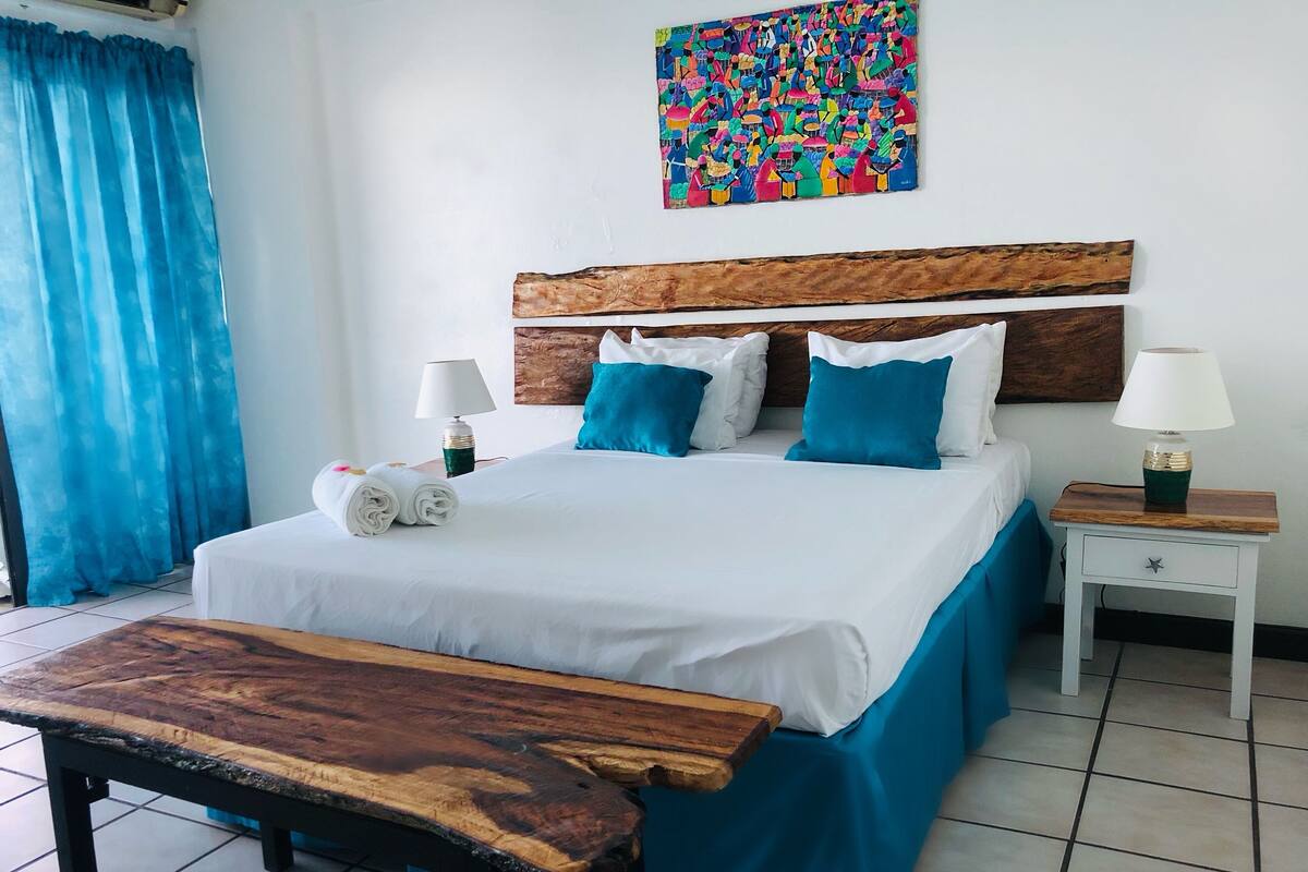 A hotel room with a bed, two bedside tables, a bench, a colorful artwork on the wall, and a window with blue curtains.
