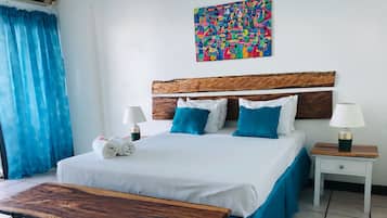 Una habitación de hotel con una cama, un banco de madera, una mesita de noche, una lámpara y un cuadro abstracto colorido en la pared.