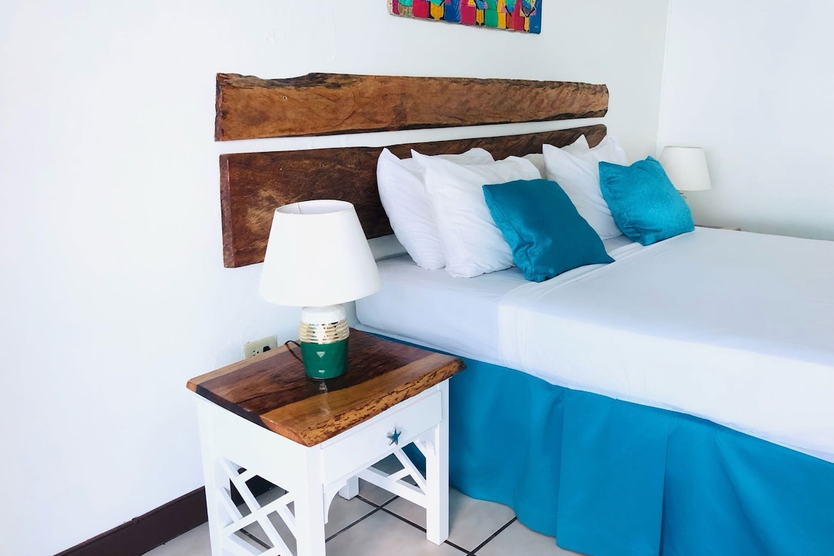 A bed with white bedding and blue curtains, a wooden nightstand with a lamp, and a colorful wall art piece above the bed.
