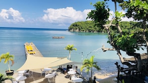 Une terrasse au bord de la plage, avec des tables et des chaises, d’où l’on peut admirer une mer calme et un bateau au loin.