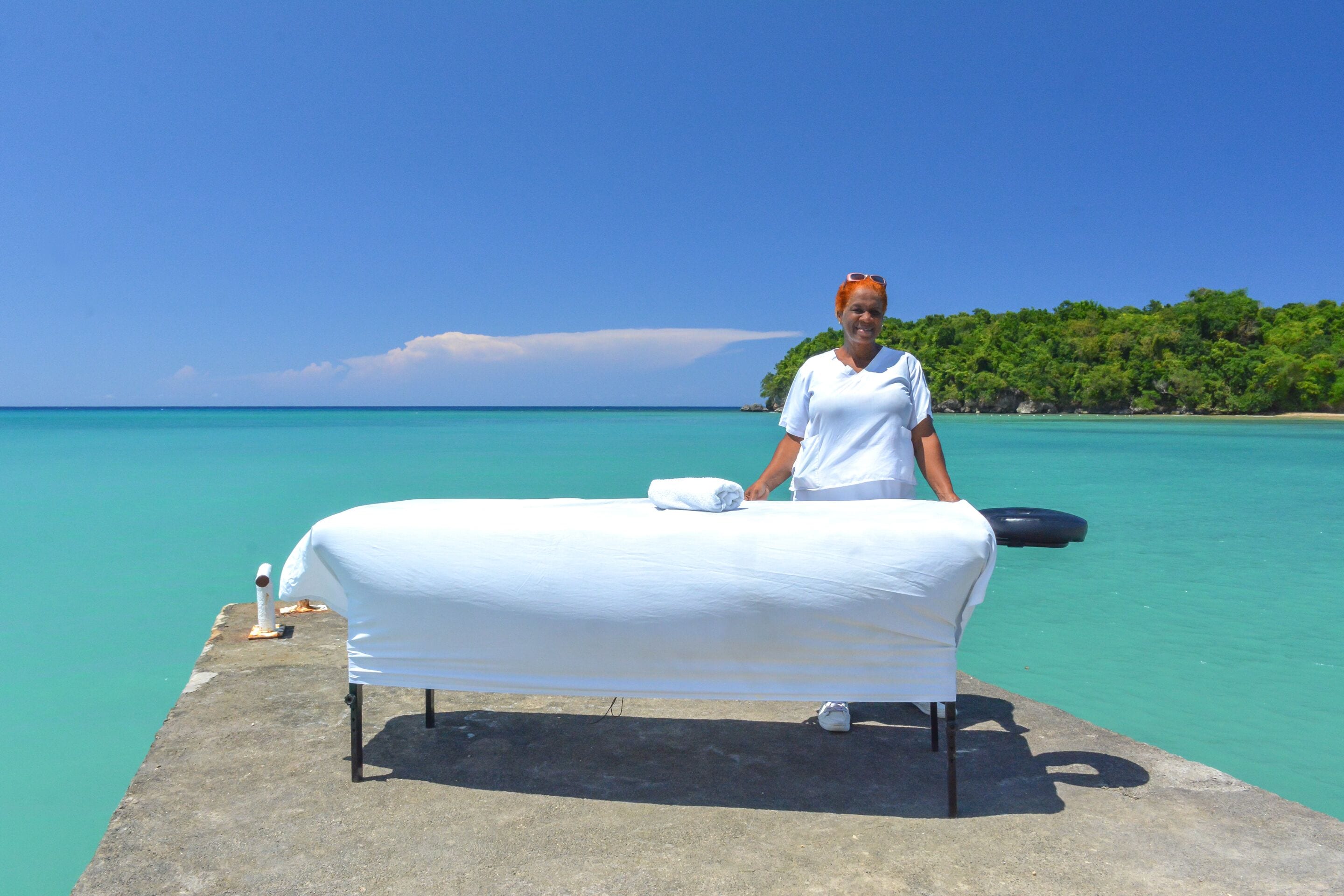A massage table set up on a concrete platform by the sea with a clear view of the turquoise water and a forested island in the background.
