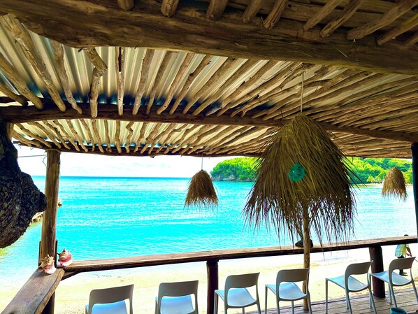 A wooden deck with chairs overlooking a turquoise sea, a thatched roof, and a view of a lush green island.