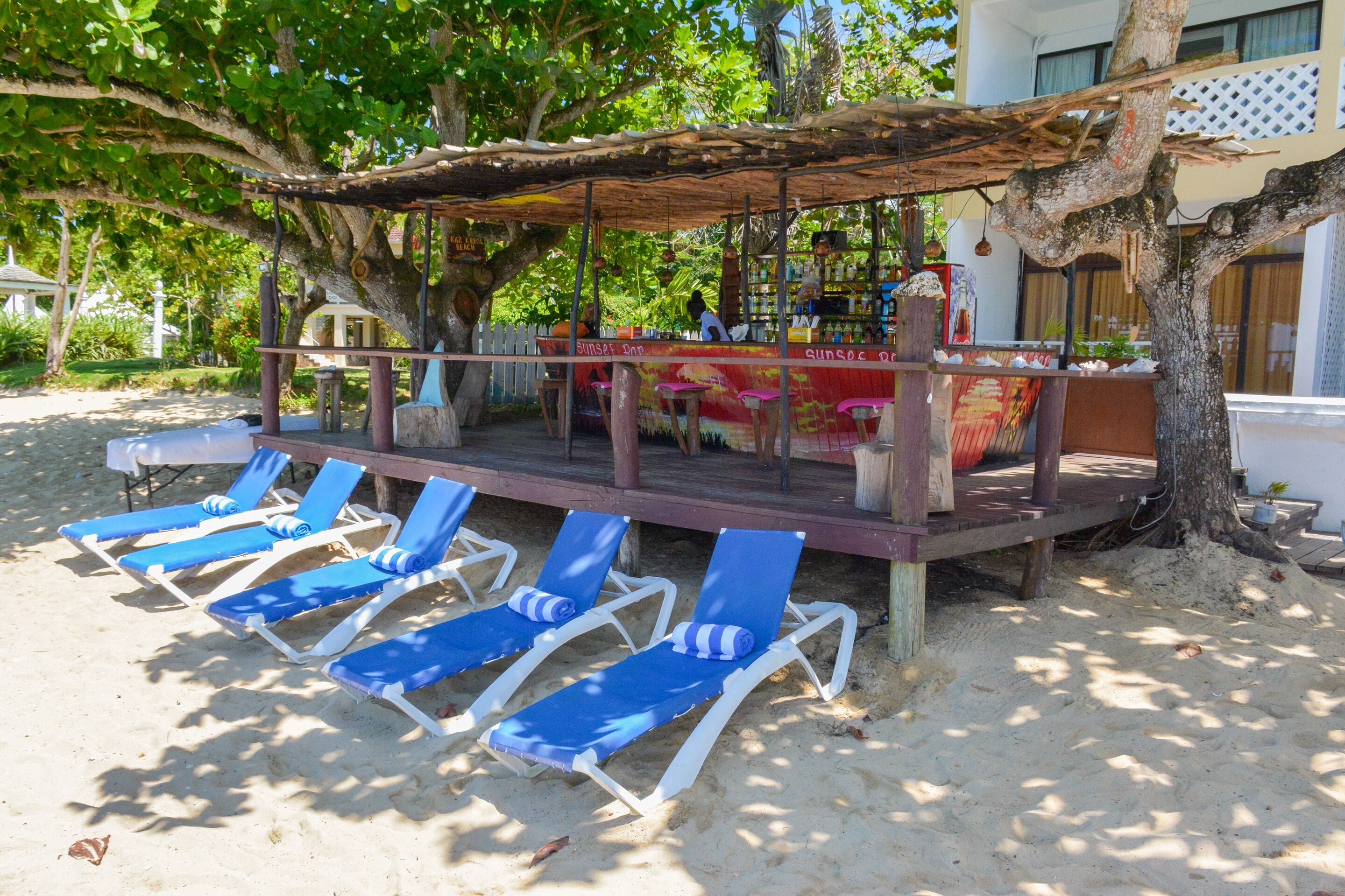 A beachside bar with a wooden deck, blue lounge chairs, and a shaded area under a tree.