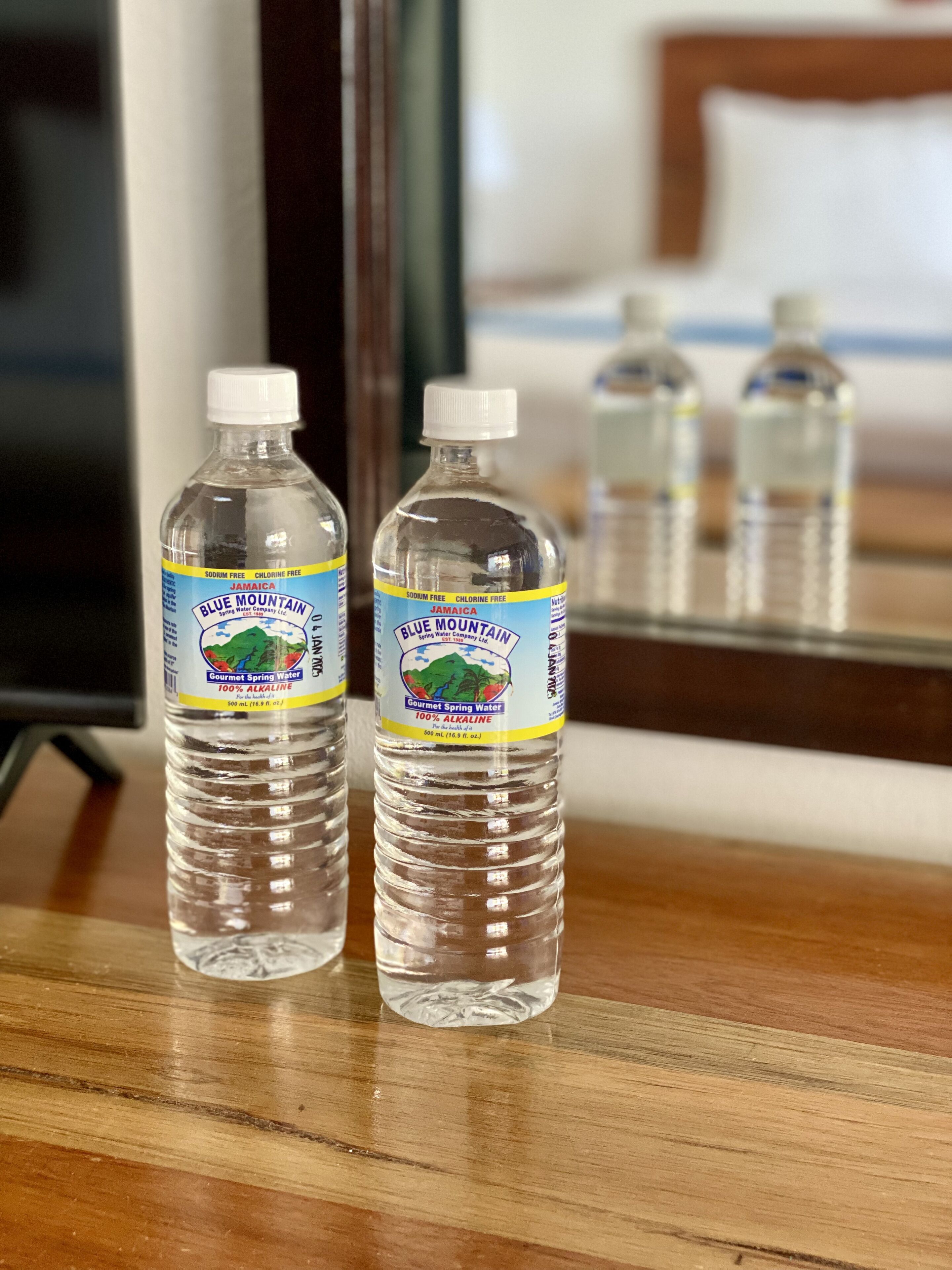 Two bottles of Jamaica Blue Mountain water on a wooden surface with a mirror reflecting a bed and a TV in the background.