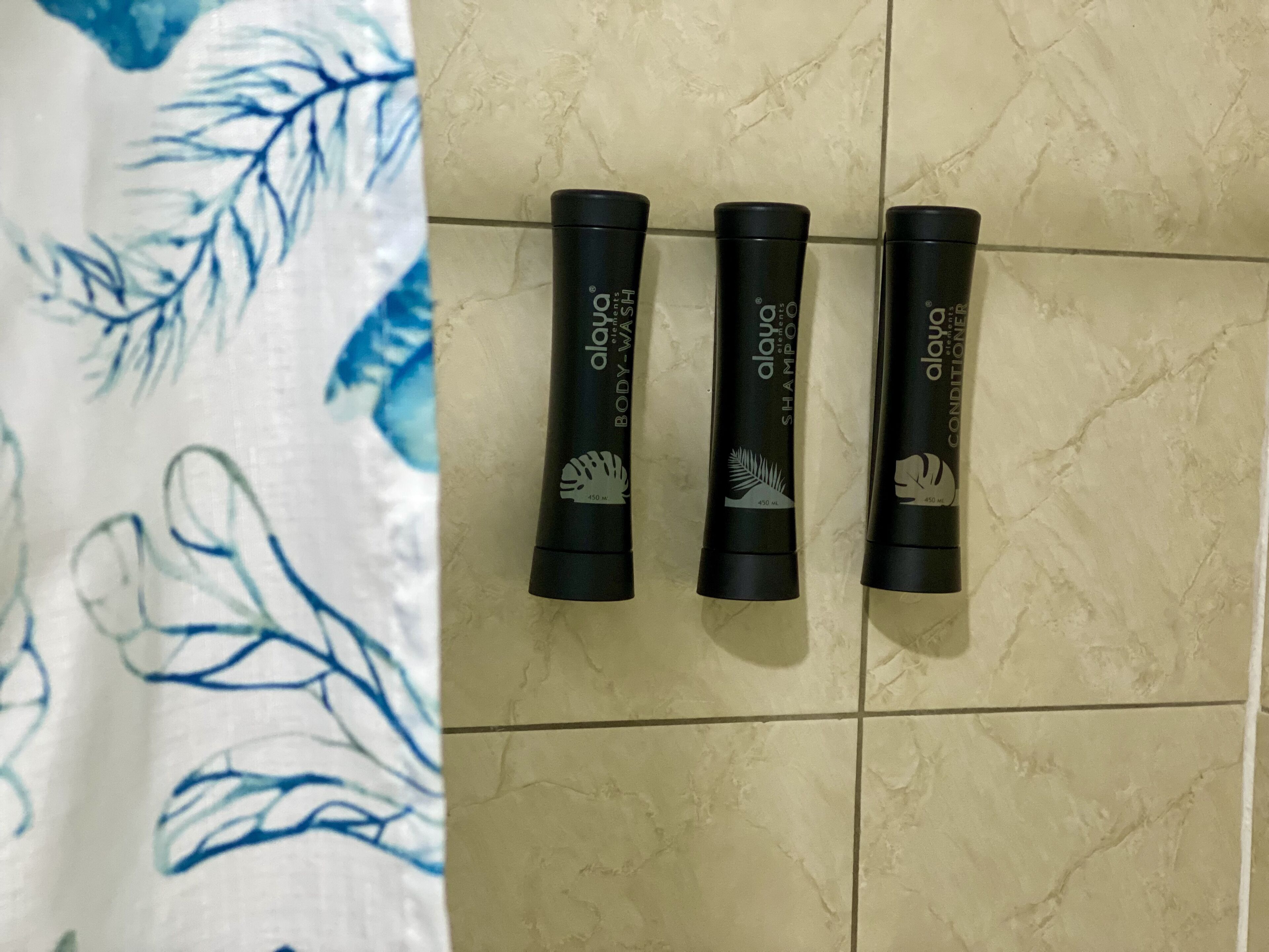 Trois flacons noirs de gel douche Alua sont posés sur un mur carrelé.