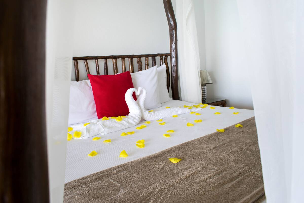 A neatly made bed with a swan-shaped towel, white and red pillows, and yellow flower petals on the bedspread.