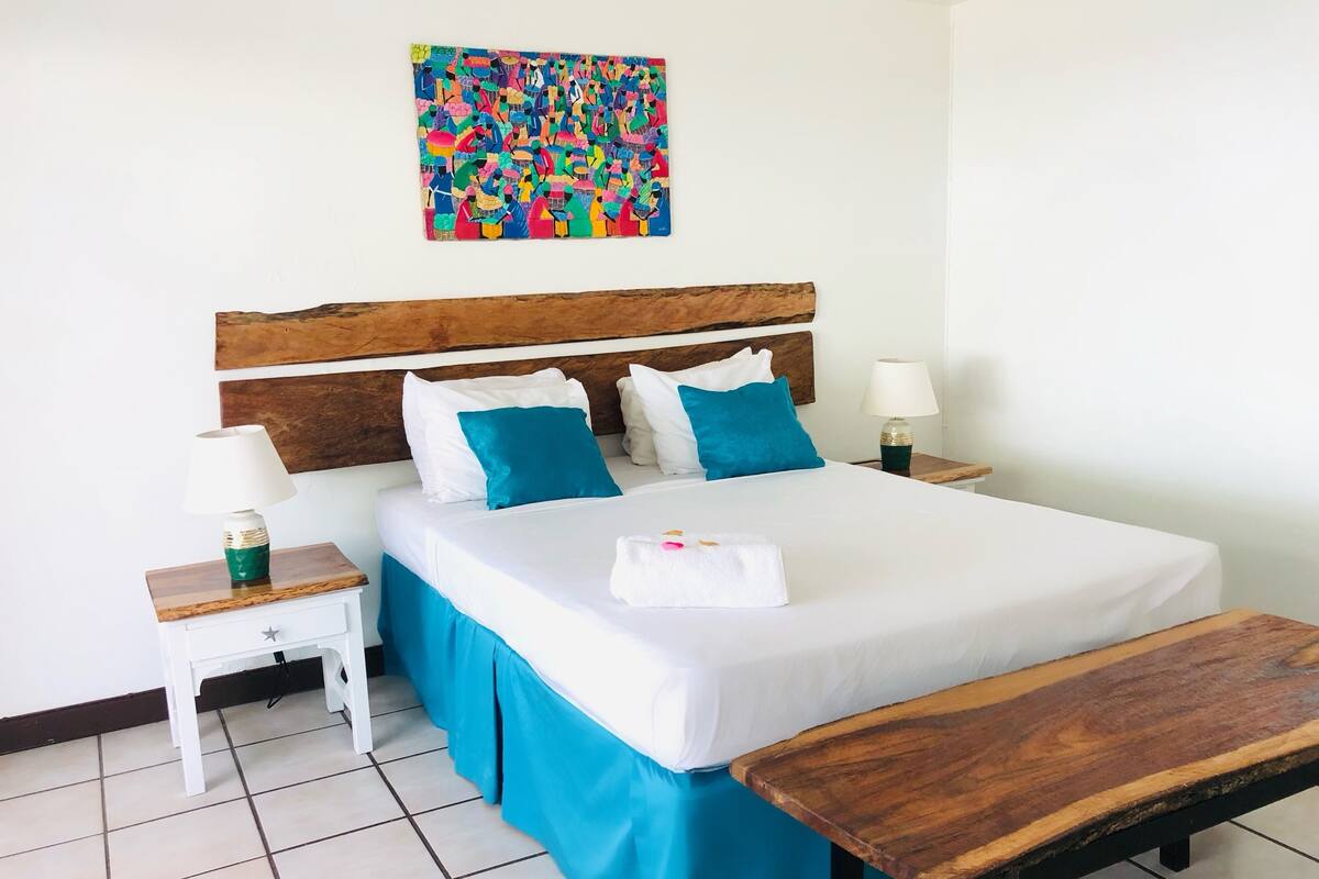 A neatly made bed with a wooden headboard, a bedside table with a lamp, a colorful abstract painting on the wall, and a wooden bench in the room.