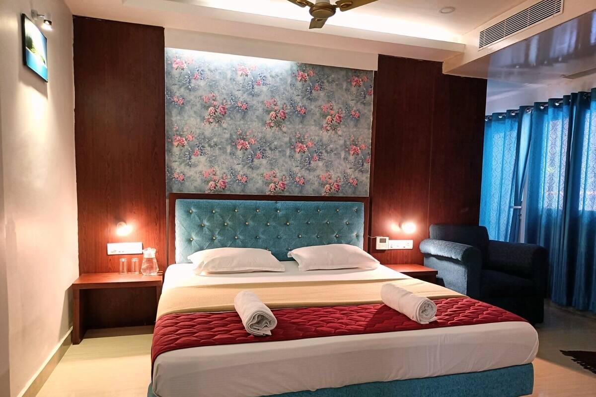 A hotel room with a large bed, a blue headboard, floral wallpaper, a blue curtain, and a nightstand with a lamp.