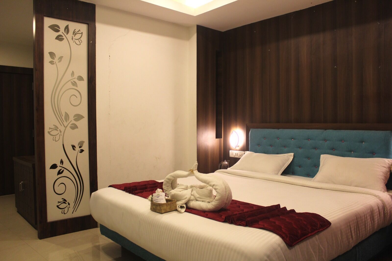 A hotel room with a large bed, a wooden headboard, a decorative wall panel, and a bedside table with a lamp.