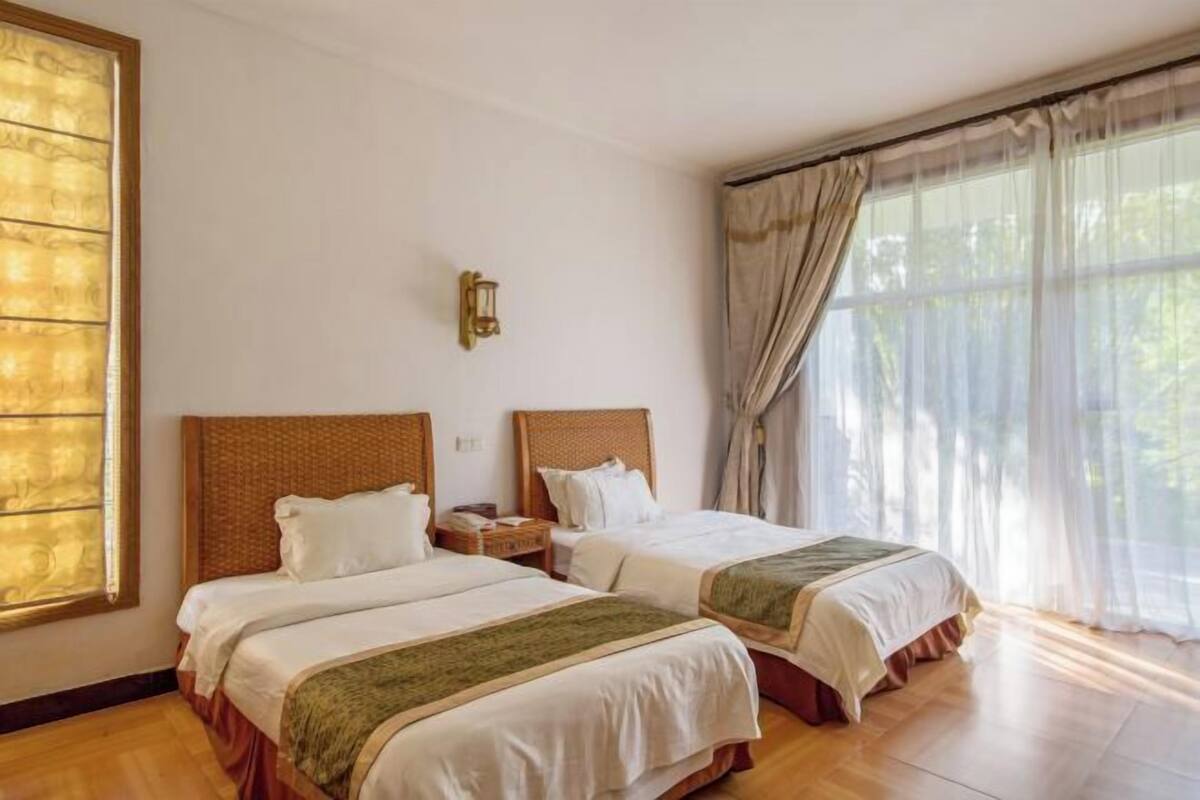 A hotel room with two beds, a nightstand, a wall-mounted lamp, and large windows with sheer curtains.
