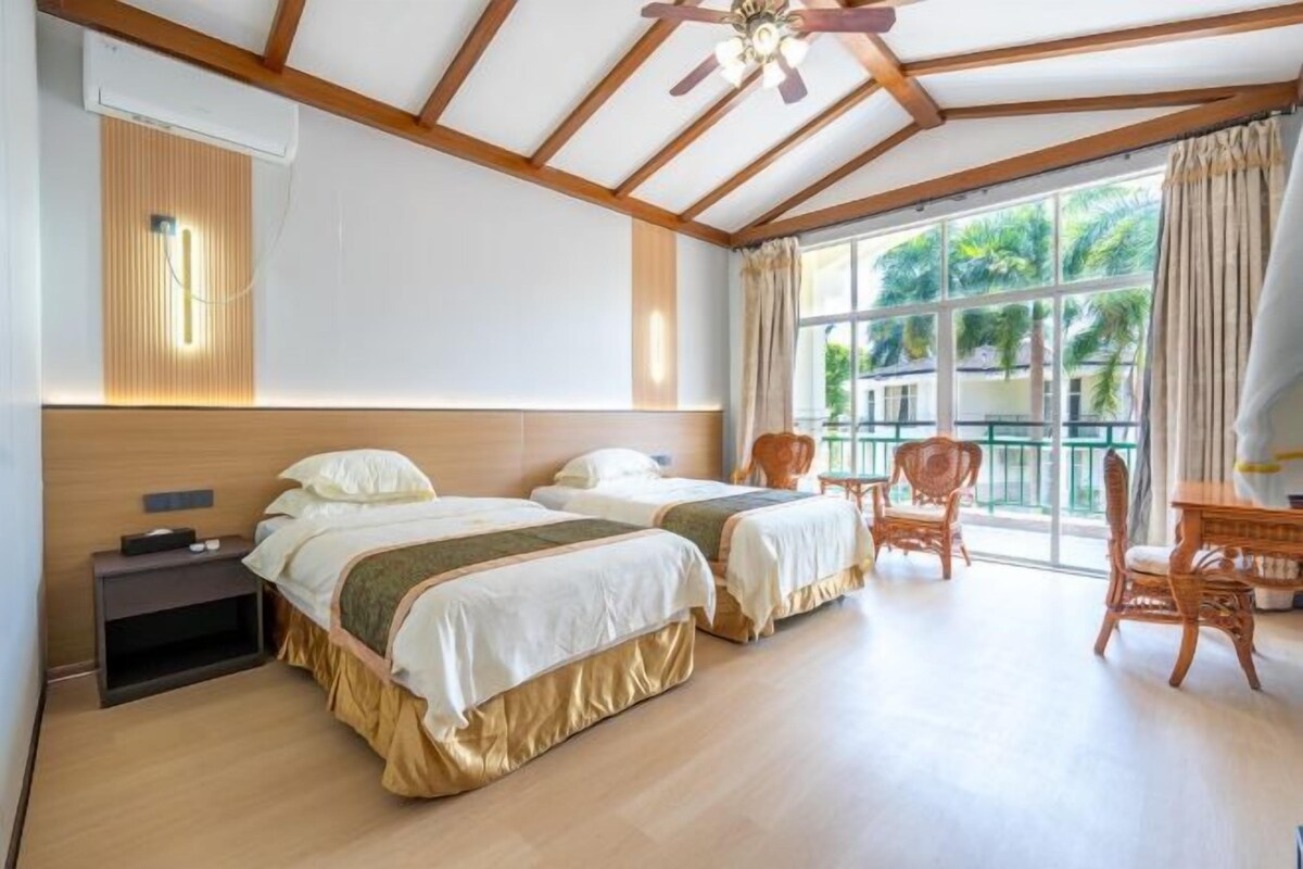 A room with two beds, a ceiling fan, a window with a view, and a chair.