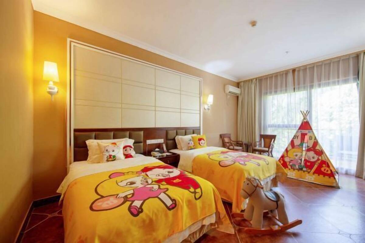 A hotel room with two beds, each with a cartoon-themed bedspread, a large window with curtains, a wooden floor, and a teepee decoration.