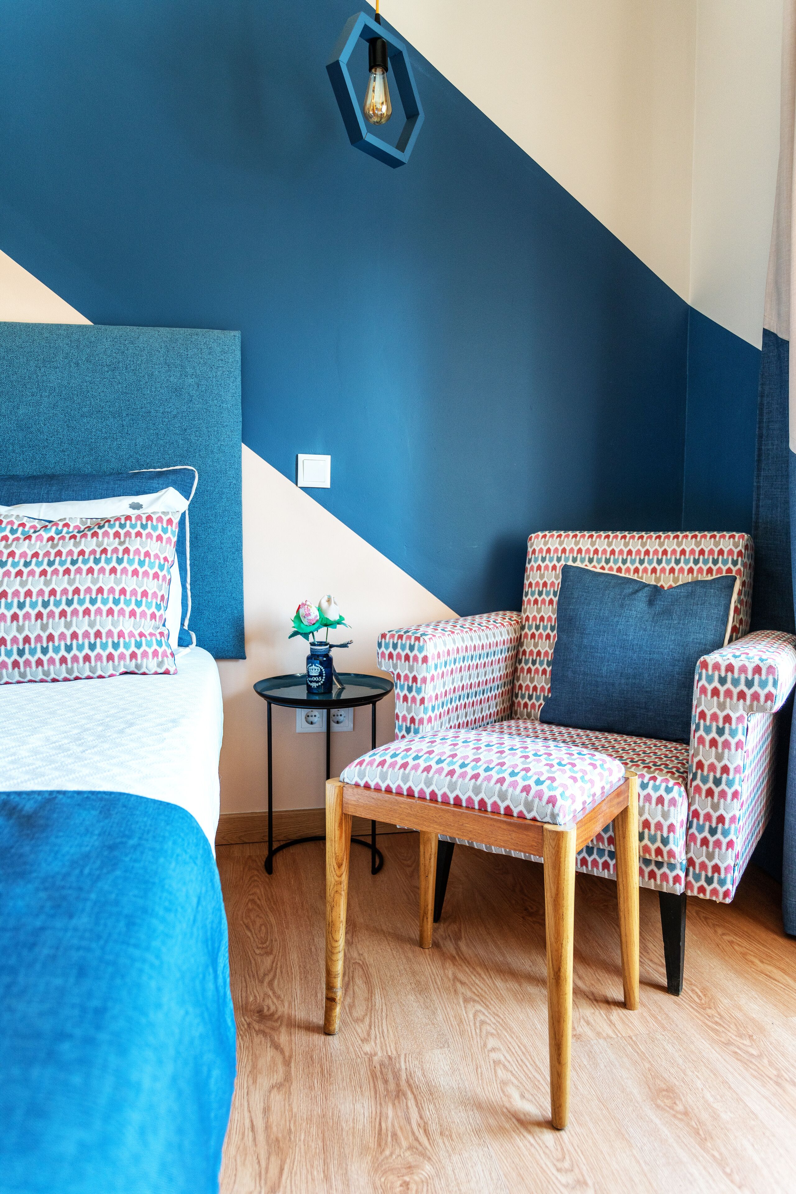 Una habitación con una cama, un sillón con estampado, una mesita con un jarrón y una lámpara hexagonal original.