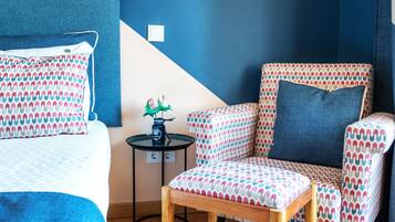 Una habitación con una cama, un sillón con estampado, una mesita con un jarrón y una lámpara hexagonal original.
