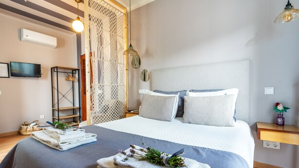 A neatly made bed with a grey and white duvet, a bedside table with a small plant, and a wall-mounted TV. - Downtown Alegria (Tavira)