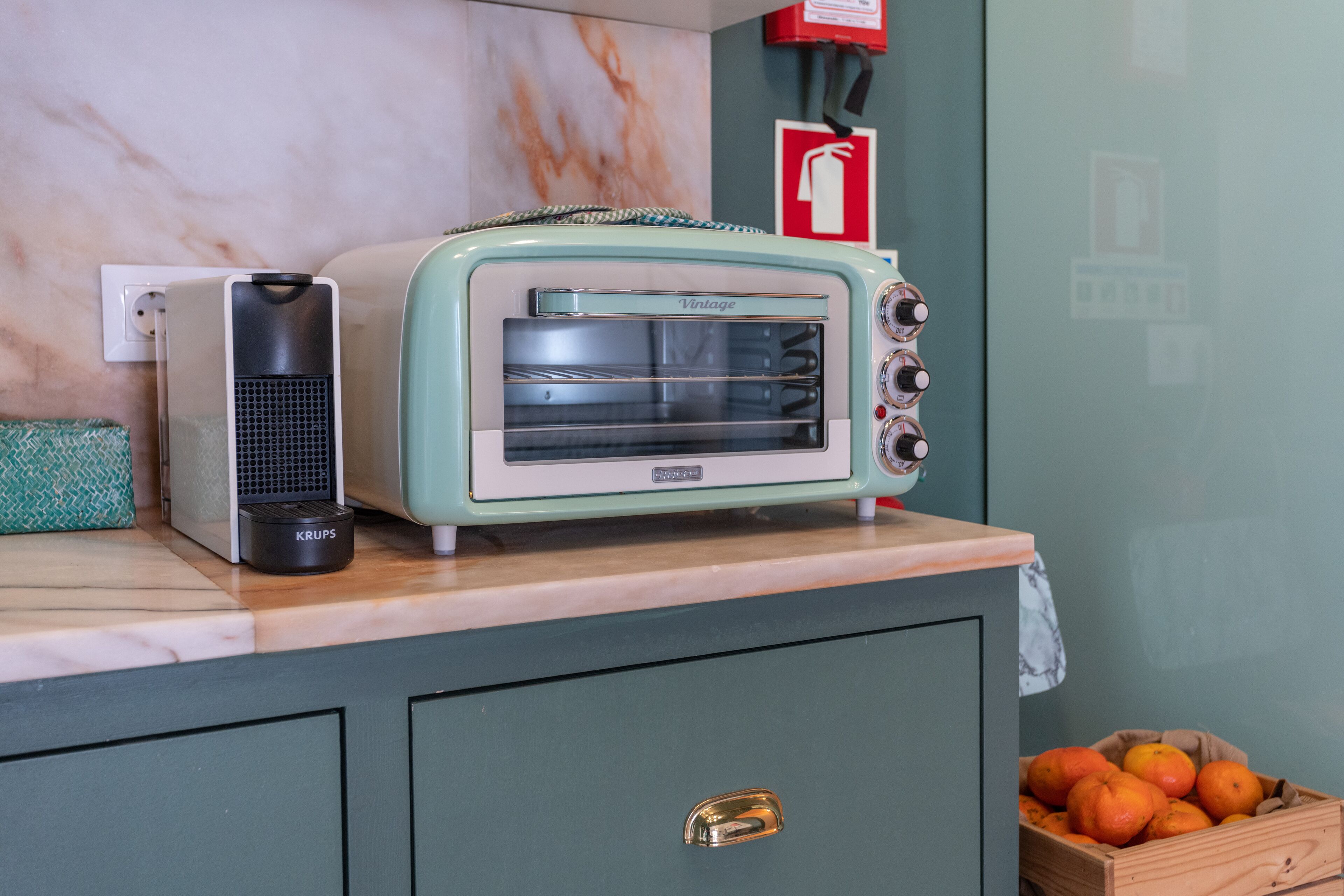 A vintage toaster oven on a kitchen counter with a marble backsplash and a wooden crate of oranges.