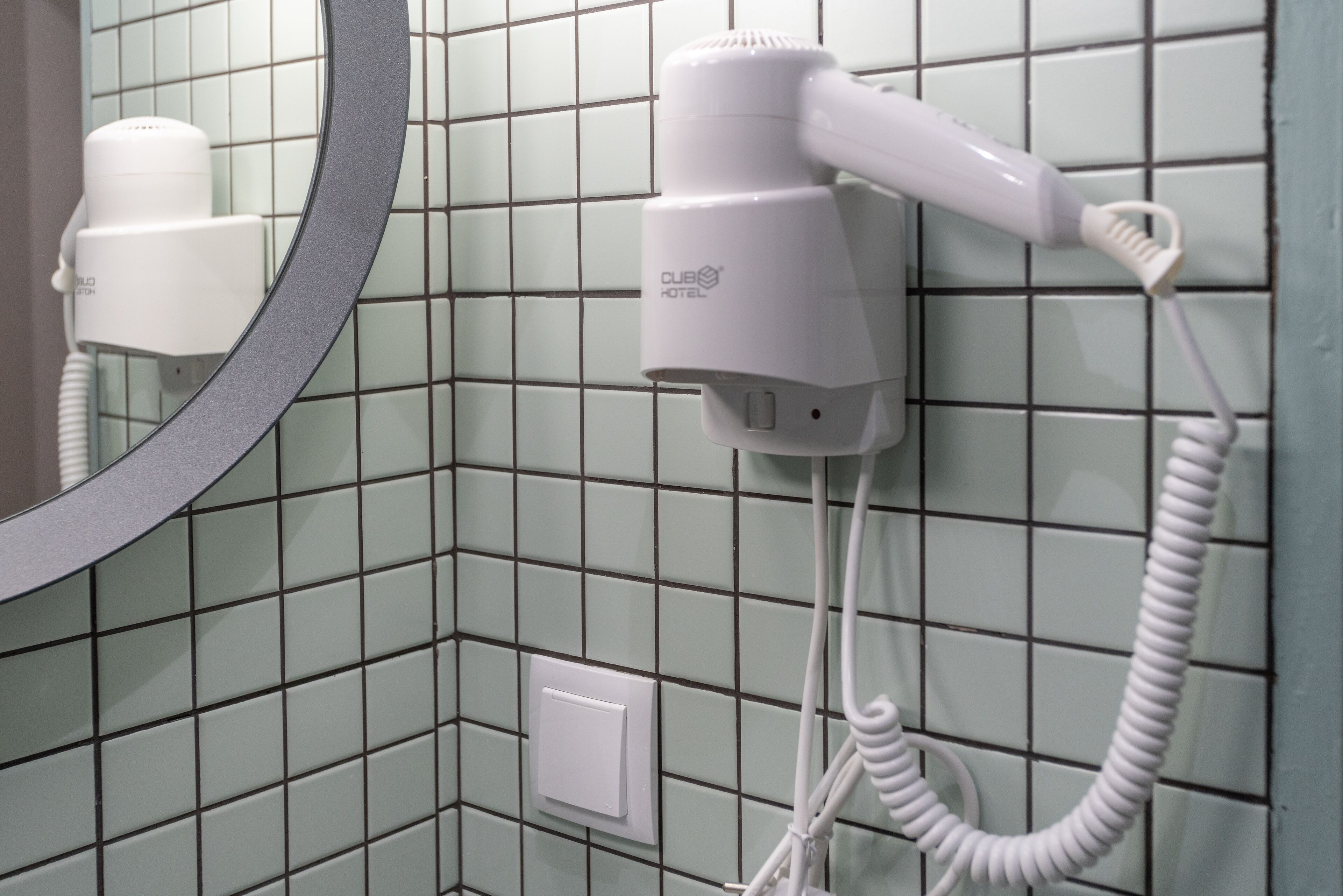 Un sèche-cheveux fixé sur un mur recouvert de carreaux, avec un miroir au-dessus.