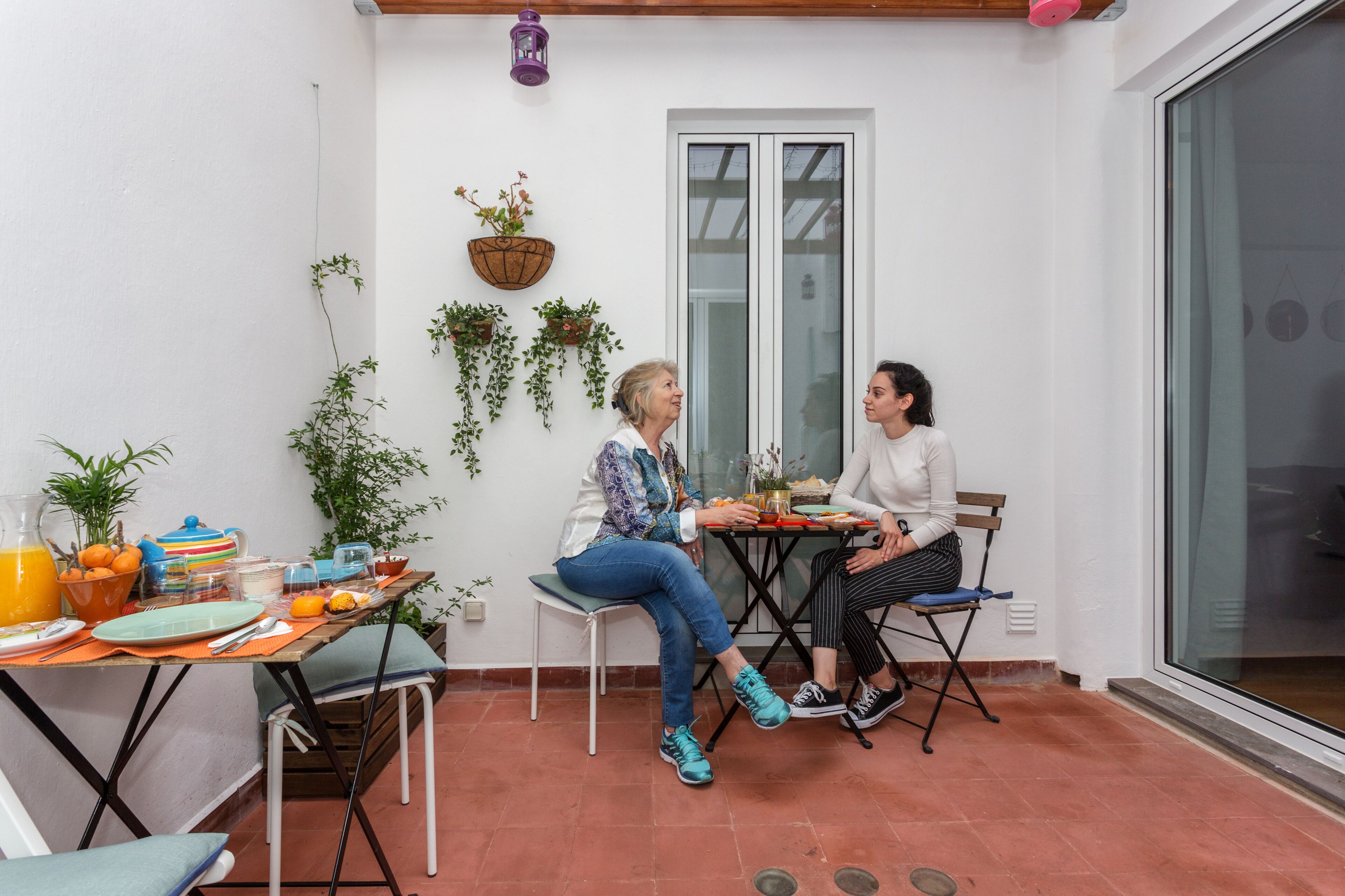 Pequeño patio amurallado con una mesa para dos personas, rodeado de macetas con plantas y una ventana.