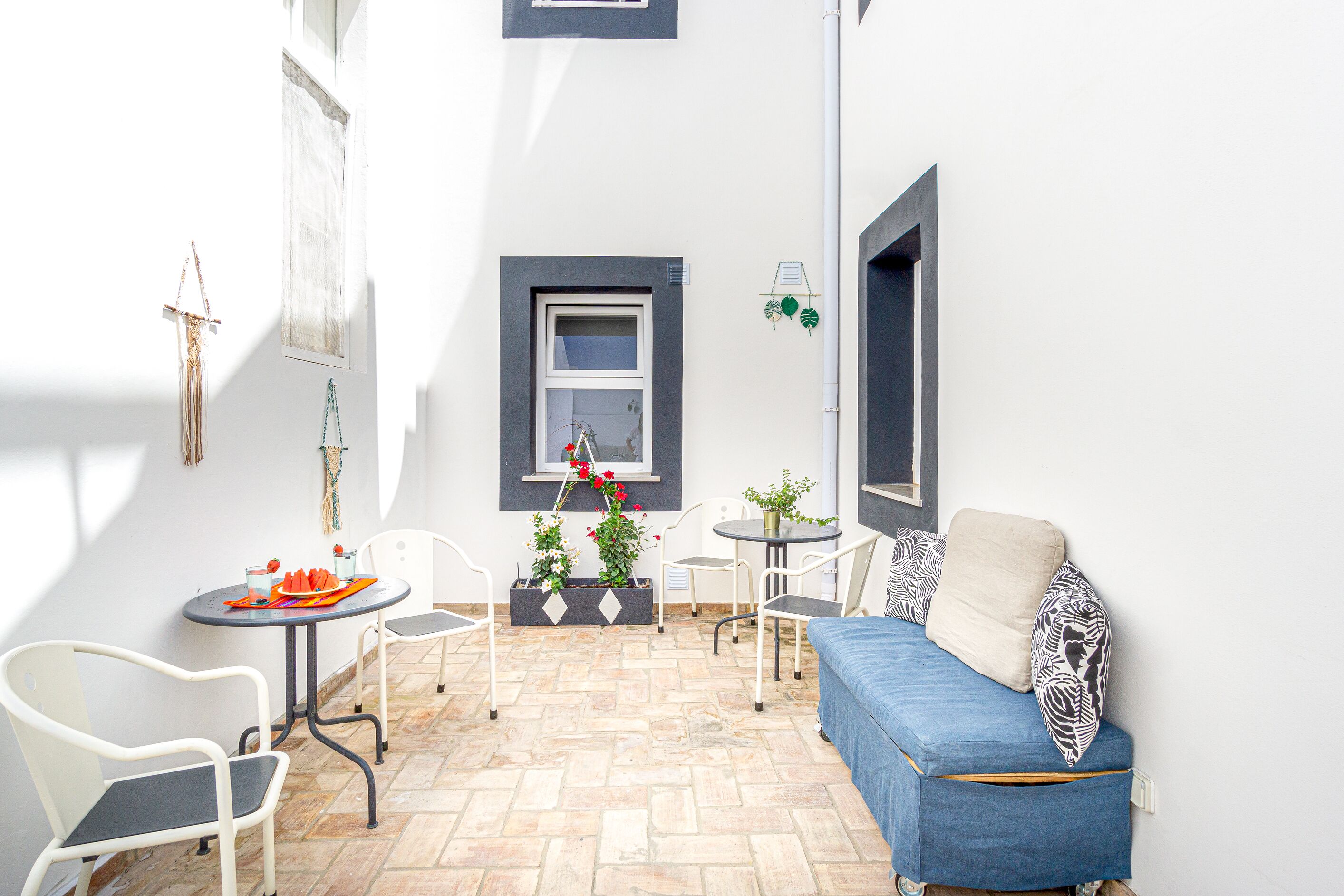 Een klein, zonnig terras met een blauwe bank, witte stoelen, een tafeltje en potplanten.