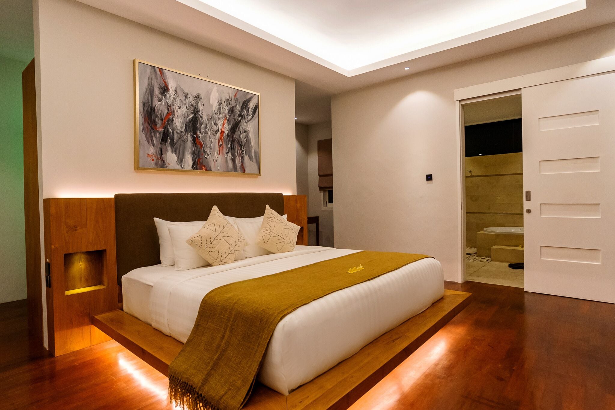 Un dormitorio moderno con una cama grande, un cabecero de madera, una mesita de noche y un cuadro abstracto de gran tamaño. Se ve un baño a través de una puerta abierta.