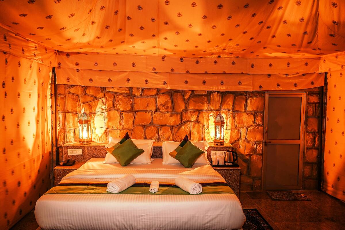 Una habitación con pared de piedra, cama con almohadas, mesita de noche, farol y una puerta.