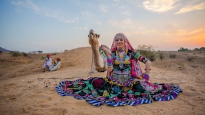 A person sitting on the sand with a camel, surrounded by a desert landscape. - Desert Over Night Camp & Resort (Jaisalmer)