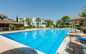 A swimming pool with clear blue water, surrounded by lounge chairs and umbrellas, with buildings in the background.