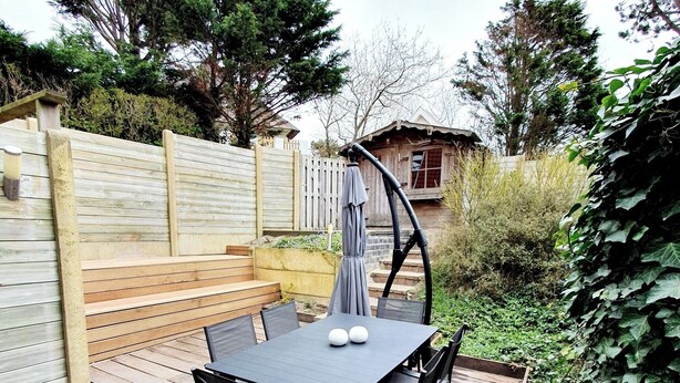 A wooden deck with a table and chairs, surrounded by a wooden fence and greenery.
