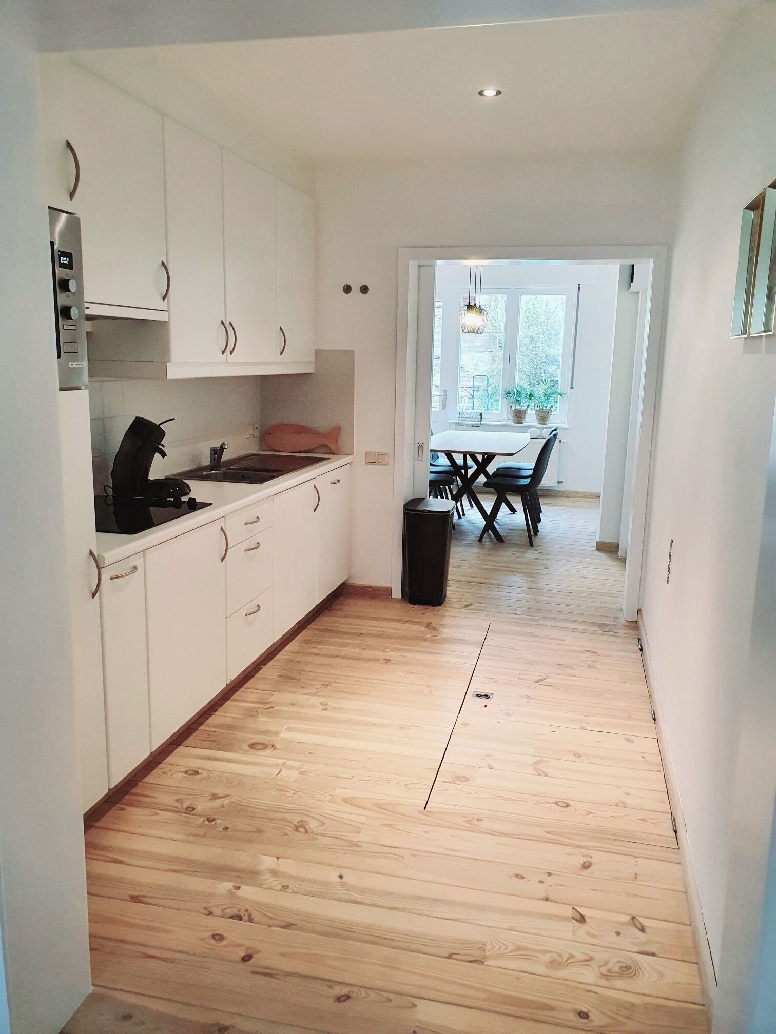 Una cocina moderna con armarios blancos, encimera negra y suelo de madera. Se distingue un área de comedor con una mesa y sillas a través de una puerta abierta.