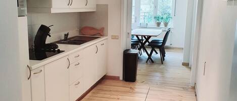 Una cocina moderna con armarios blancos, encimera negra y suelo de madera. Se distingue un área de comedor con una mesa y sillas a través de una puerta abierta.