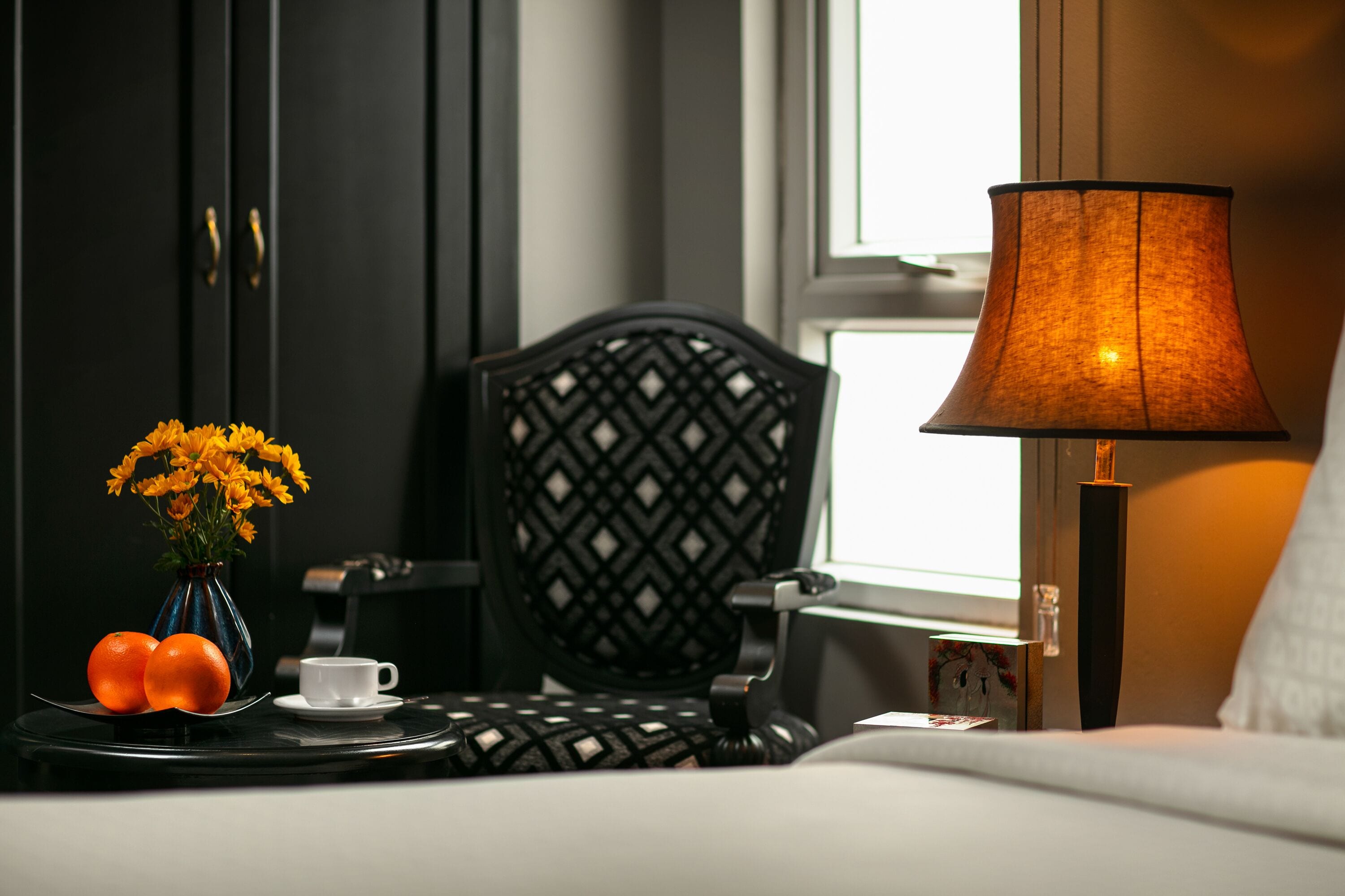 A neatly arranged hotel room with a black chair, a bedside table with a lamp, a vase with flowers, and a plate with oranges.