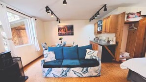 Una habitació acollidora amb un sofà blau estampat, sòl de fusta, zona de cuina i un llit amb edredó blanc.