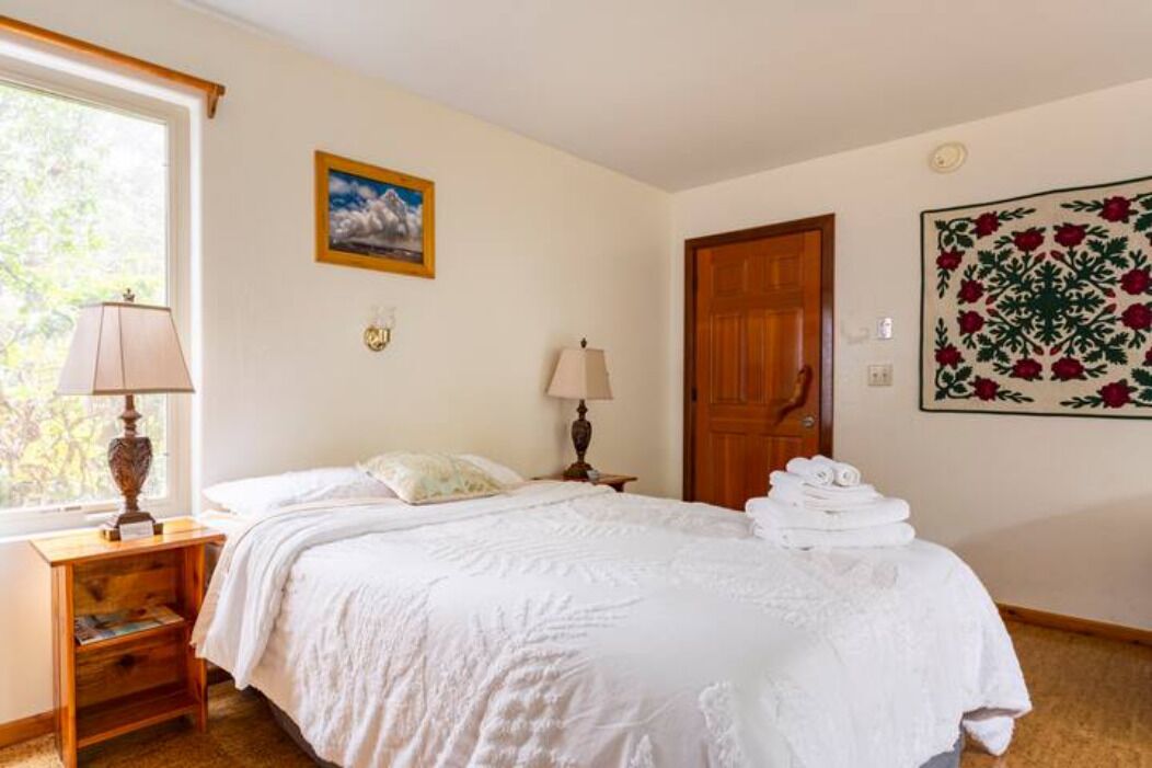 A bedroom with a bed, two bedside lamps, a wooden nightstand, a window with a view, a door, and a wall with a floral tapestry.