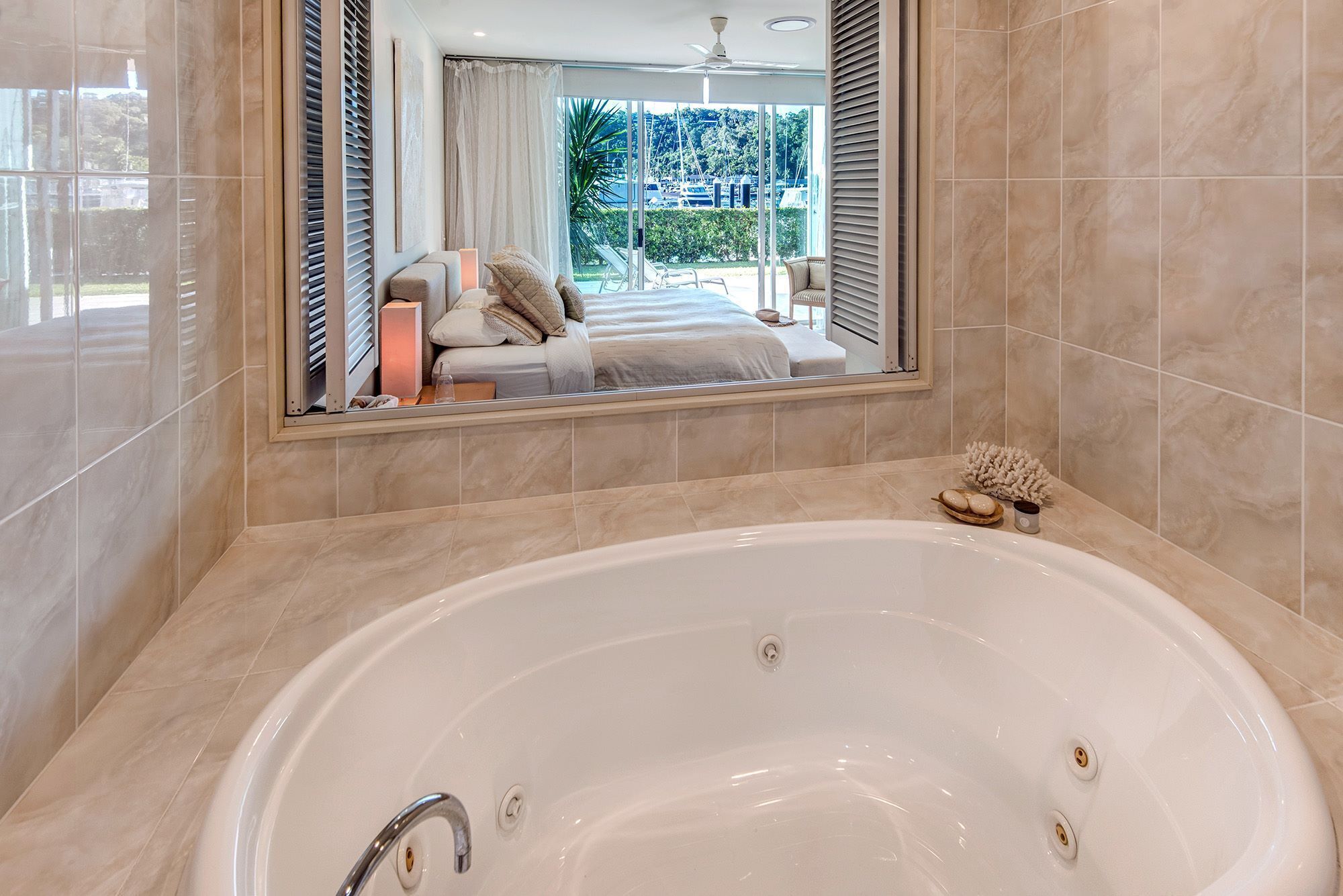A bathroom with a large bathtub, a mirror reflecting a bedroom with a bed and curtains, and a view of a marina outside.