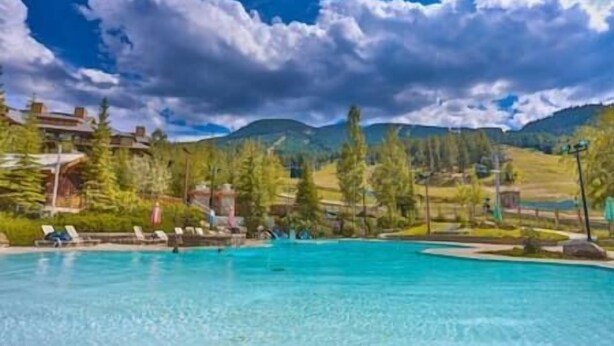 A clear blue pool with lounge chairs, surrounded by trees and a mountainous landscape.