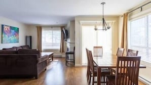 A spacious living area with a sofa, dining table, and chairs. There is a painting on the wall and a window providing natural light.