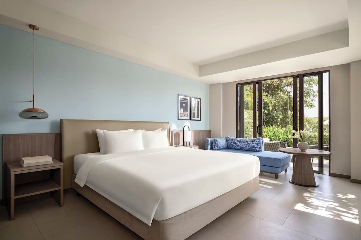 Habitación de hotel moderna con una cama grande, una mesita de noche, una pequeña mesa con un florero, un sofá azul y ventanas amplias que ofrecen vistas a zonas verdes.