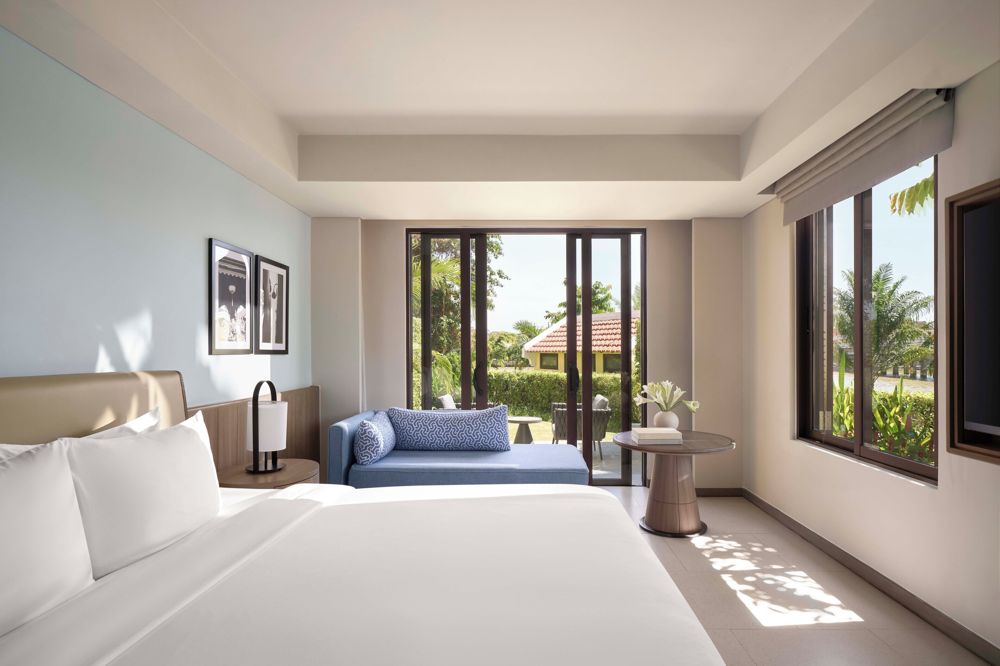 A modern hotel room with a large bed, a sofa, a small table, and a view of the outdoors.
