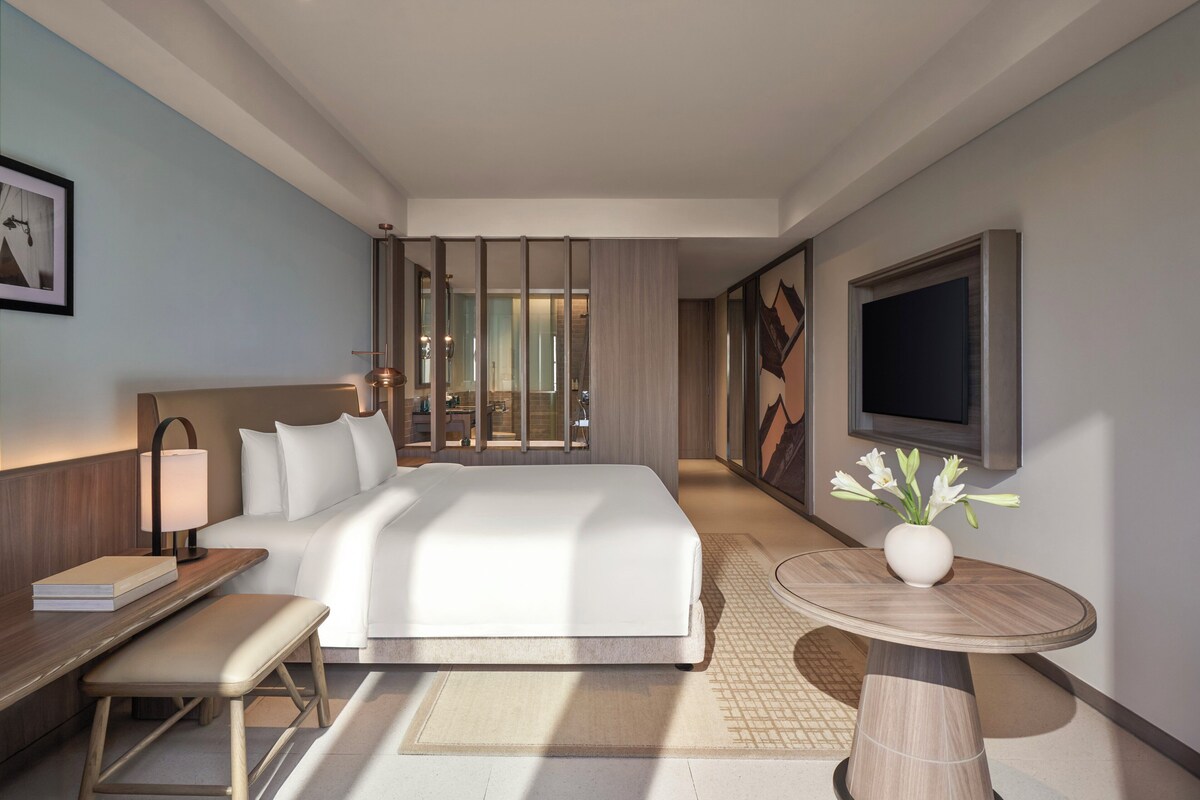 A modern hotel room with a large bed, bedside table, lamp, and a round table with a vase of flowers.