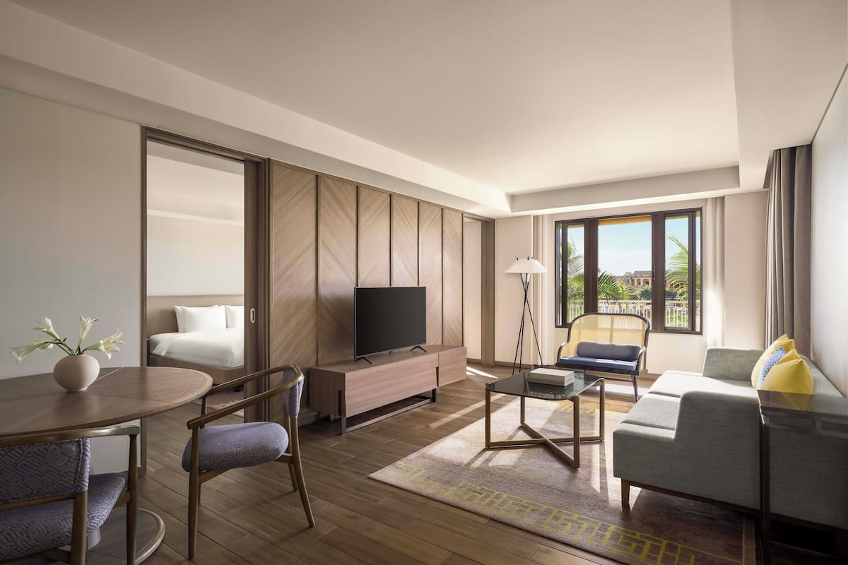 Una habitación de hotel moderna con suelo de madera, un televisor de pantalla plana sobre un mueble de madera, un sofá, una mesa de comedor con sillas y un amplio ventanal con vistas a palmeras.